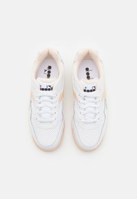 White sneakers with perforated texture, beige accents, and white laces. Features a rounded toe and a soft, cushioned interior.