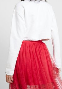 Vit kortärmad sweatshirt med långa ärmar, ihopparad med en voluminös röd tyllkjol som har en texturerad lagerdesign.