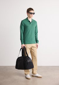 Cămașă polo verde cu mânecă lungă, pantaloni bej, adidași albi și o geantă de voiaj mare, neagră, cu bretele pentru umăr. Persoană purtând ochelari de soare.