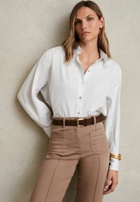 Cămașă albă cu nasturi, cu mâneci lungi, combinată cu pantaloni mulți de culoare maro deschis și o curea maro. Țesătură netedă, design structurat, detalii minime.
