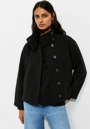 Donna con capelli scuri che indossa orecchini a cerchio, una giacca nera abbottonata con collo alto, un top nero e jeans azzurri, in piedi su sfondo bianco.