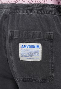 Temno siva jeans tkanina z teksturirano površino. Ima elastičen pas in belo oznako prišito na zadnjem žepu z modrim besedilom.