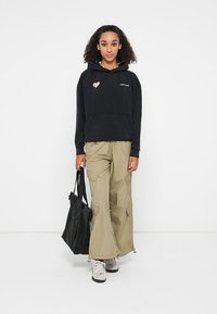 Zwarte hoodie met hartlogo, gecombineerd met losse kaki cargobroek met zakken. Model houdt een zwarte tote bag vast en draagt sneakers.