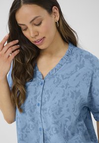 Svijetloplava košulja kratkih rukava s teksturiranim cvjetnim uzorkom, volanastim dekolteom u obliku V i dva gumba sprijeda.