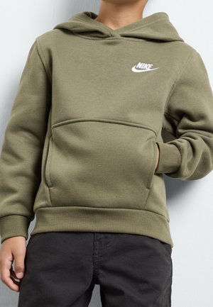 Enfant portant un sweat à capuche Nike vert olive avec poche avant et pantalon noir, debout contre un mur gris clair.