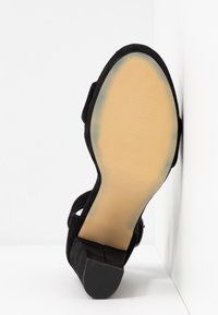 Chaussure à talon haut en daim noir vue de dos, présentant une semelle lisse avec une zone antidérapante texturée et une bride incurvée à la cheville.