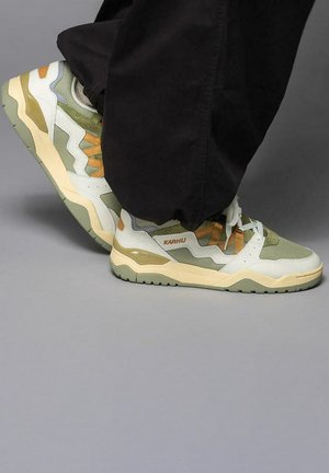 Persona con zapatillas Karhu en verde oliva y beige, con cordones blancos y pantalones negros, de pie sobre un suelo gris.