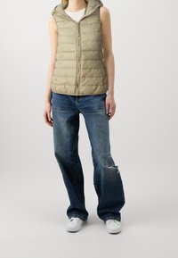 Ljus khaki ärmlös puffer väst med huva, dragkedjestängning och kviltad textur; kombinerad med slitna blå vida jeans och vita sneakers.