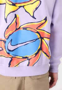 Lila sweatshirt med en stor, färgglad grafisk soldesign med livfulla orange, gula och blå accenter. Nike-logotyp ingår.