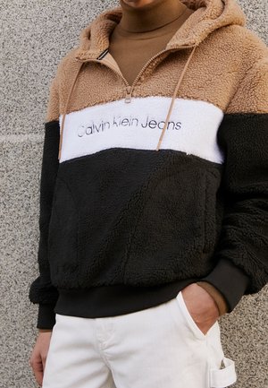 Osoba ubrana w polarowy sweter w kolorach brązowym, białym i czarnym z napisem „Calvin Klein Jeans”, w połączeniu z białymi spodniami, ręka w kieszeni.