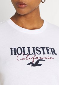 Bílá bavlněná tričko s výšivkou "HOLLISTER California" v námořnické modři a červené, s silueta loga pod textem. Krátké rukávy.