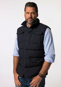 Svart vadderad väst med hög krage, framfickor och knapprading, bärs över en ljusblå skjorta och denimjeans.