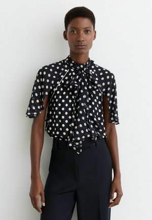 Femeie purtând o bluză neagră cu buline albe și o fundă mare la guler, asortată cu pantaloni negri cu talie înaltă.