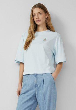 Młoda kobieta z długimi włosami, ubrana w jasnoniebieski oversize T-shirt z małym kwiatowym haftem i jasnoniebieskie jeansy z wysokim stanem.