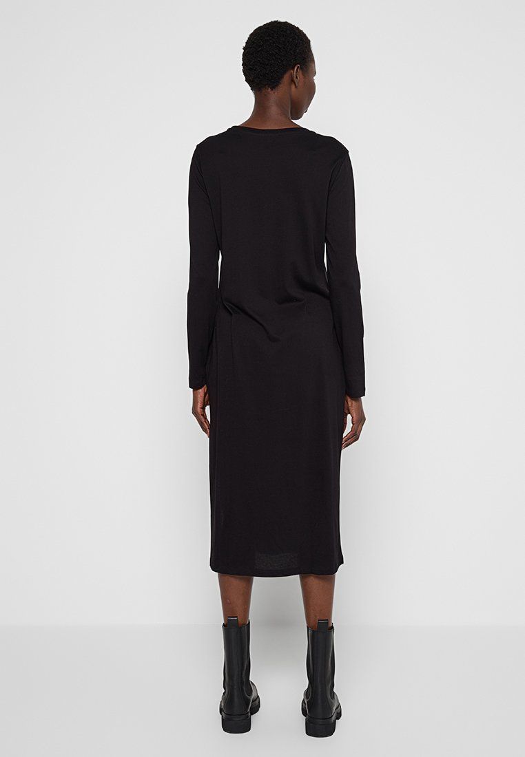 Persona vestita con un abito midi nero a maniche lunghe e stivali neri alla caviglia, in piedi di spalle contro uno sfondo bianco semplice.