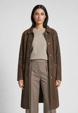 Kvinde iført en brun knælang frakkemodel med knapper over en lys beige sweater og taupe højtaljede plisserede bukser står mod en grå baggrund.