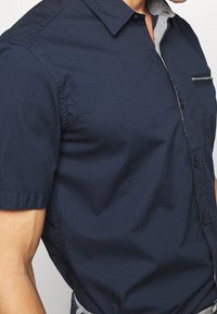 Košile s krátkým rukávem v námořnické modři s vertikálními pruhy, s šedým límcem a náprsní kapsou s dekoračním akcentem. Texturovaná tkanina.