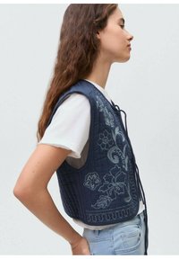 Quiltana mornarica brezrokavnik s cvetličnim vezenjem, z vezalnim sprednjim delom in brezrokavno zasnovo, oblečen preko belega srajce in svetlo modrih kavbojk.