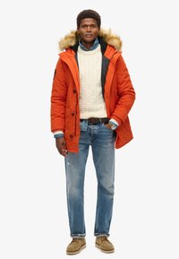Parka naranja con capucha forrada de piel, forro interior negro, combinada con un suéter de punto en color crema, camisa de botones azul, vaqueros claros y zapatos beiges.