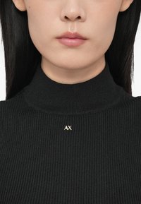 Egy nő alsó arca és nyaka fekete bordázott garbós pulóverben, amelynek gallérján egy apró fém „AX” logó található.