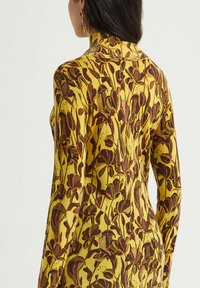 Top a maniche lunghe in tessuto giallo con stampa floreale in marrone scuro e bordeaux. Design aderente con collo alto e texture liscia.