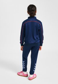 Marineblaue Trainingsjacke mit roten Akzenten, ausgestattet mit einem Reißverschlusskragen und gemusterten Streifen an den Hosen. Kombiniert mit rosa Turnschuhen und weißen Socken.