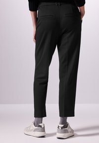 Pantalon noir avec une coupe ajustée, doté de poches latérales, d'une texture lisse et d'une longueur raccourcie, associé à des baskets grises et blanches.