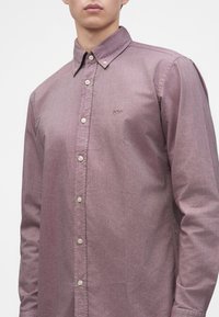 Homme portant une chemise à manches longues mauve discret avec un col boutonné et un petit logo brodé sur la poitrine.