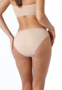 Donna che indossa biancheria intima senza cuciture color beige e reggiseno, vista da dietro su sfondo bianco.