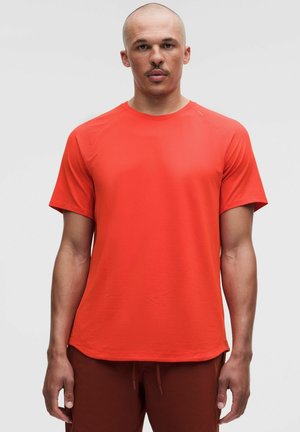 Mann mit rasiertem Kopf, der ein hellrotes kurzärmeliges Sportshirt und dunkelrote Shorts trägt, blickt nach vorne vor einem schlichten Hintergrund.