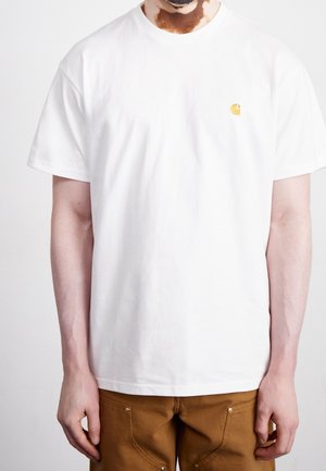 Homme atteint de vitiligo portant un t-shirt blanc uni à manches courtes avec un petit logo jaune et un pantalon marron sur fond blanc.