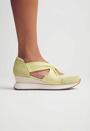 Pie con zapato de cuña de color verde claro con tiras de satén cruzadas y puntera de malla sobre un fondo blanco liso.