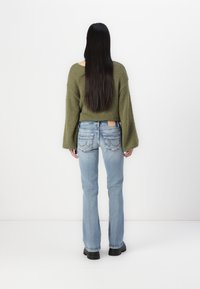 Olivgrüner, oversize Pullover mit weiten Ärmeln, kombiniert mit hellblauen, weit geschnittenen Jeans, die eine Applikation auf der Gesäßtasche haben, sowie schwarzen Stiefeln.