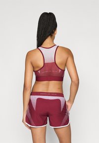 Tumši sarkans sporta krūšturis ar tīkls muguru, kopā ar atbilstošiem tumši sarkaniem šortiem ar baltu apmali un rakstainiem izšuvumiem. Apģērbam redzams teksts.