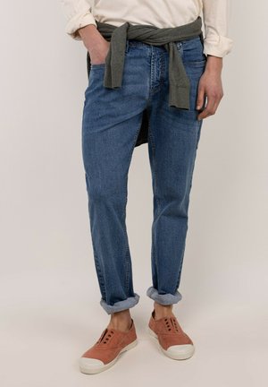Persona con vaqueros azules remangados, camisa blanca con las mangas remangadas, suéter gris atado alrededor de la cintura y zapatos rosa sin cordones.