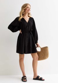 Svart linneklänning med v-ringning, knäppning fram och knytbälte. Kombineras med svarta sandaler och en flätad stråväska. Modellen står framför en neutral bakgrund.