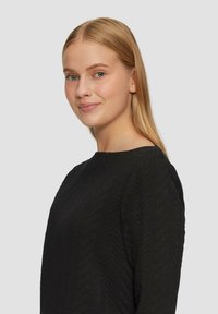 Maglione nero con texture caratterizzato da un motivo a diamante, scollatura tonda e maniche lunghe. Luce sottile e materiale morbido ben visibili.