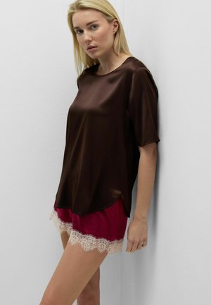 Donna bionda con top corto in raso marrone scuro e pantaloncini fucsia con bordo in pizzo bianco, appoggiata a un muro bianco.