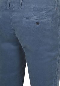 Achteraanzicht van blauwe ribfluwelen broek met focus op de rechterachterzak met een zwarte knoopsluiting.