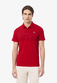 Röd polo skjorta med krage, med en geometrisk mönsterdesign i en mörkare nyans av röd. Inkluderar en grön logotyp på vänster bröst.