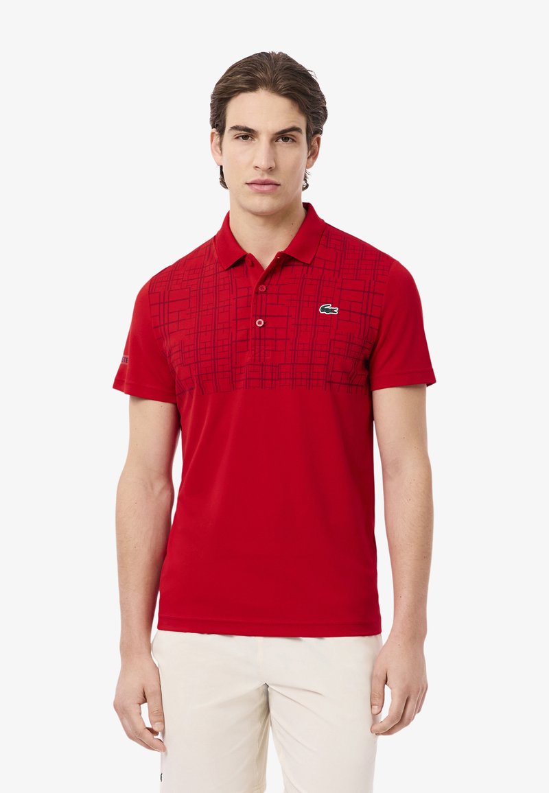 Röd polo skjorta med krage, med en geometrisk mönsterdesign i en mörkare nyans av röd. Inkluderar en grön logotyp på vänster bröst.