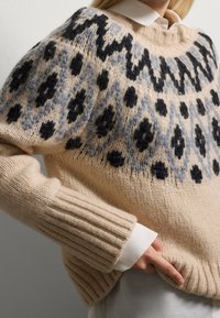 Beige Strickpullover mit schwarzen und grauen geometrischen Mustern. Gerippte Bündchen und Saum, mit rundem Ausschnitt und lässiger Passform. Weiche Textur.