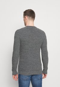Grå stickad tröja med en texturerad yta, rund halsringning och långa ärmar. Tillsammans med blå jeans, sedd bakifrån.