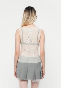 Top senza maniche in leggero beige trasparente con ricami floreali e paillettes, abbinato a una gonna grigia plissettata. Dettagli in pizzo visibili sotto.