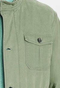 Chaqueta verde claro con un bolsillo en el pecho abotonado sobre un suéter texturizado turquesa, mostrando los ojales a lo largo de la abertura frontal de la chaqueta.