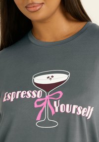 Szara koszulka z grafiką kieliszka koktajlowego z ziarnami kawy, z napisem "Espresso Yourself" w różowym kolorze.