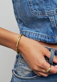 Guldfärgad metallarmband med en slät och texturerad design som bärs på en handled, tillsammans med en matchande tunn guldring, mot denimtyg.