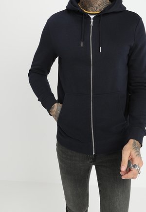 Mężczyzna w czarnej bluzie na zamek z przednimi kieszeniami i ciemnoszarych dżinsach, częściowo widoczne tatuaże na szyi i dłoniach.