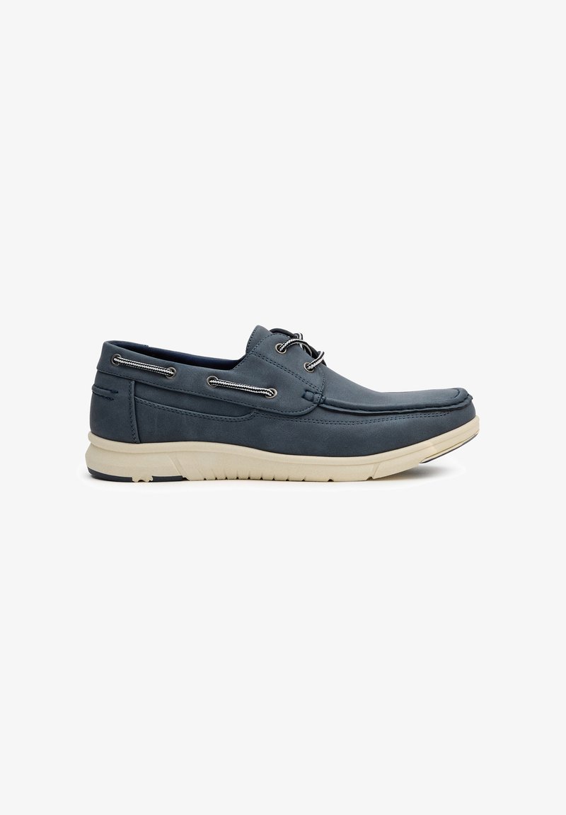 Zapatos de barco de gamuza azul marino con suela blanca, que cuentan con ojales y cordones plateados, textura suave y un diseño de punta rectangular.
