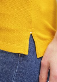 Gul stickad topp med texturerad yta, med en sidospalt och vit sömnad, kombinerad med mörk denimjeans.